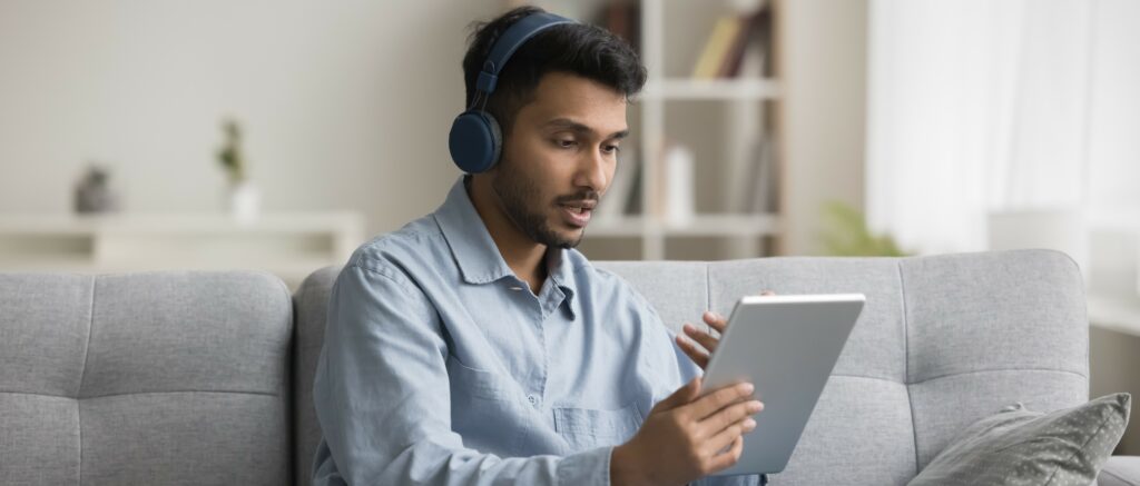 A young man sits on his couch wearing headphones and taking a video call on his tablet