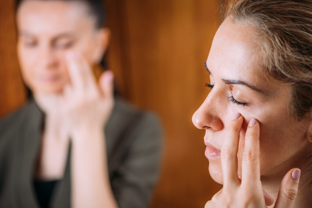 Close-up of a young woman's face. Her eyes are closed as she taps her own cheek with two fingers.