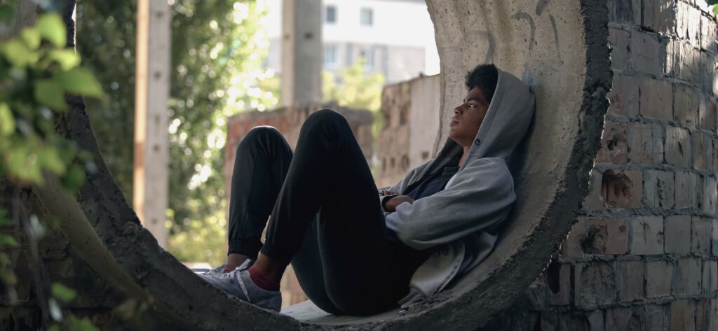 A teenage boy sits in the curve of a stone wall looking lonely and thoughtful