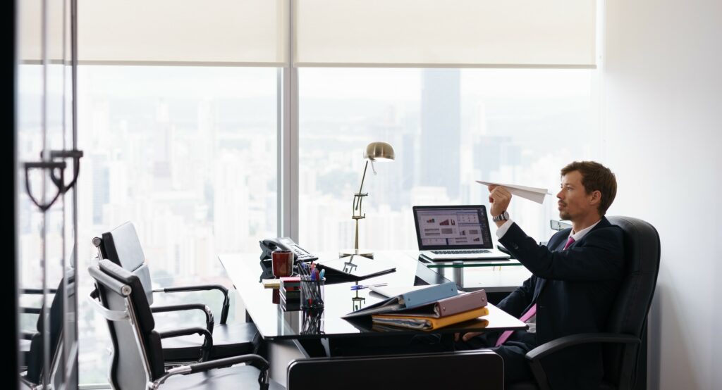 A man sits at his desk ignoring the computer and playing with a paper airplane