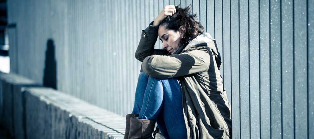 A woman sits on the edge of a low wall with one arm wrapped around her knees and the other holding her hair as she closes her eyes