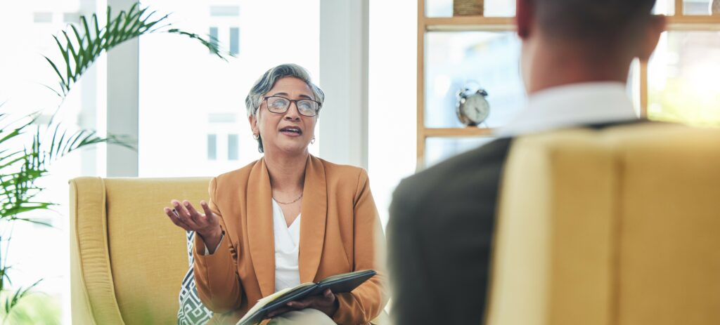 An older, female therapist and a male client sit facing each other and talking