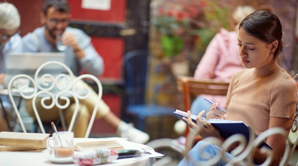 A young woman sits at a table in a cafe writing in her journal