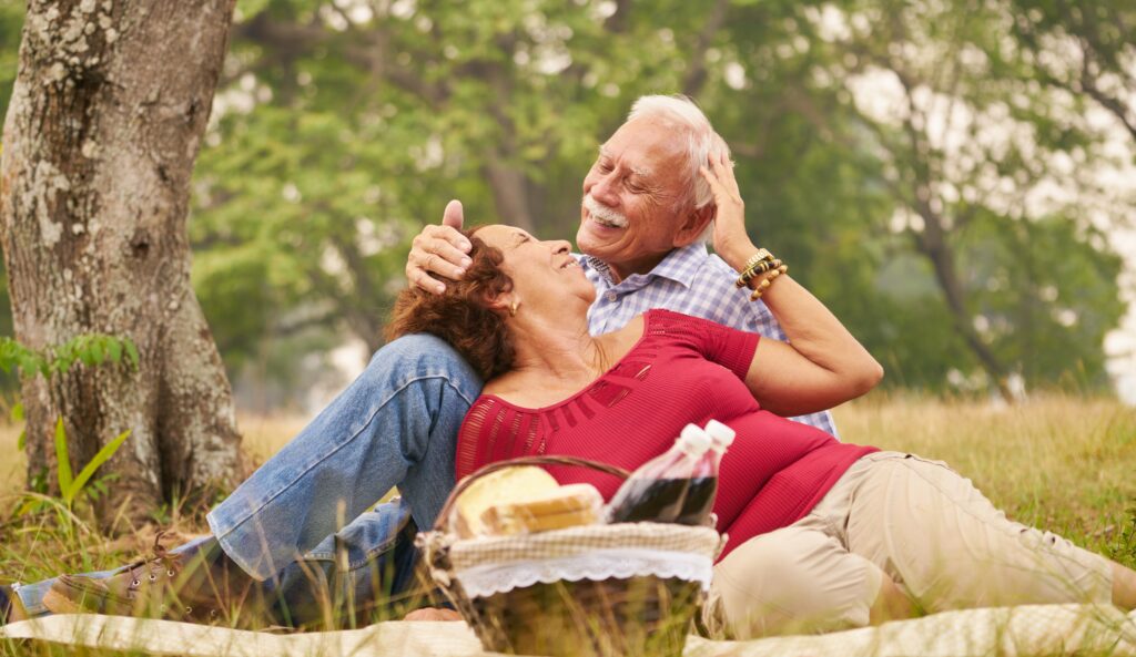 An older couple looks at each other and smiles as they eat a picnic in front of a tree