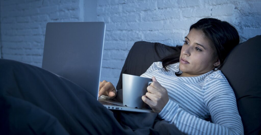 A woman lays in bed scrolling on her laptop and drinking coffee