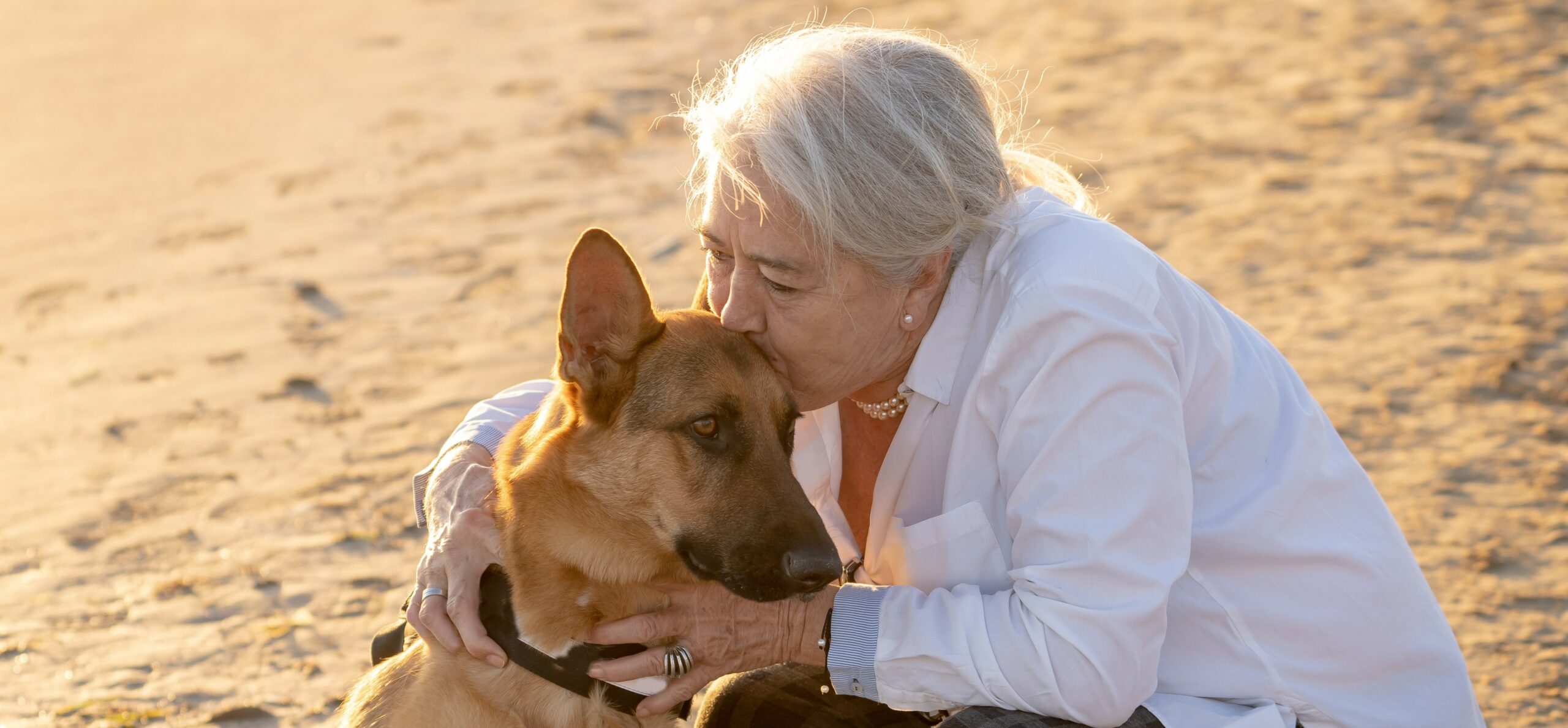 A woman leans down and kisses her dog on the forehead as they walk on the beach