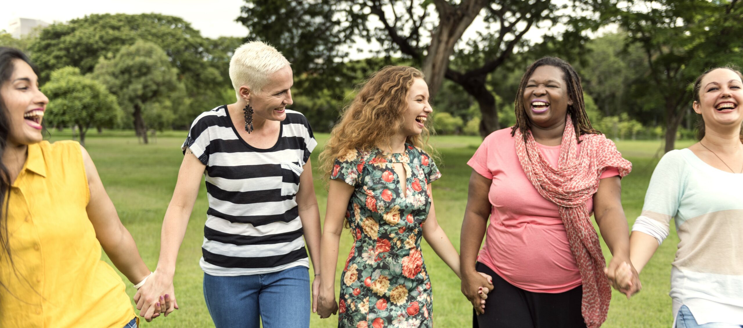 A group of women walk through a park holding hands and laughing