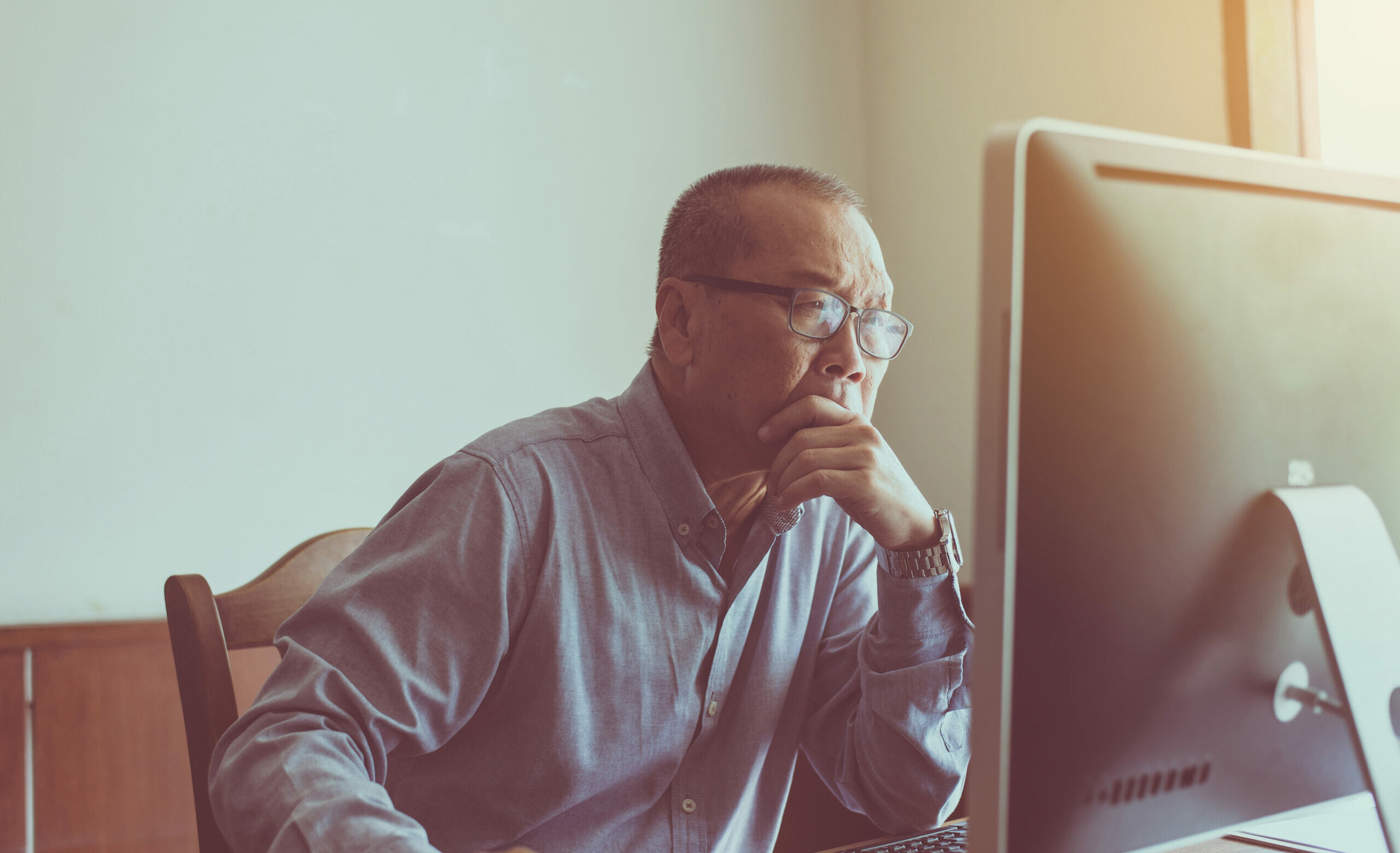Senior Asian American man looks thoughtfully at his computer monitor