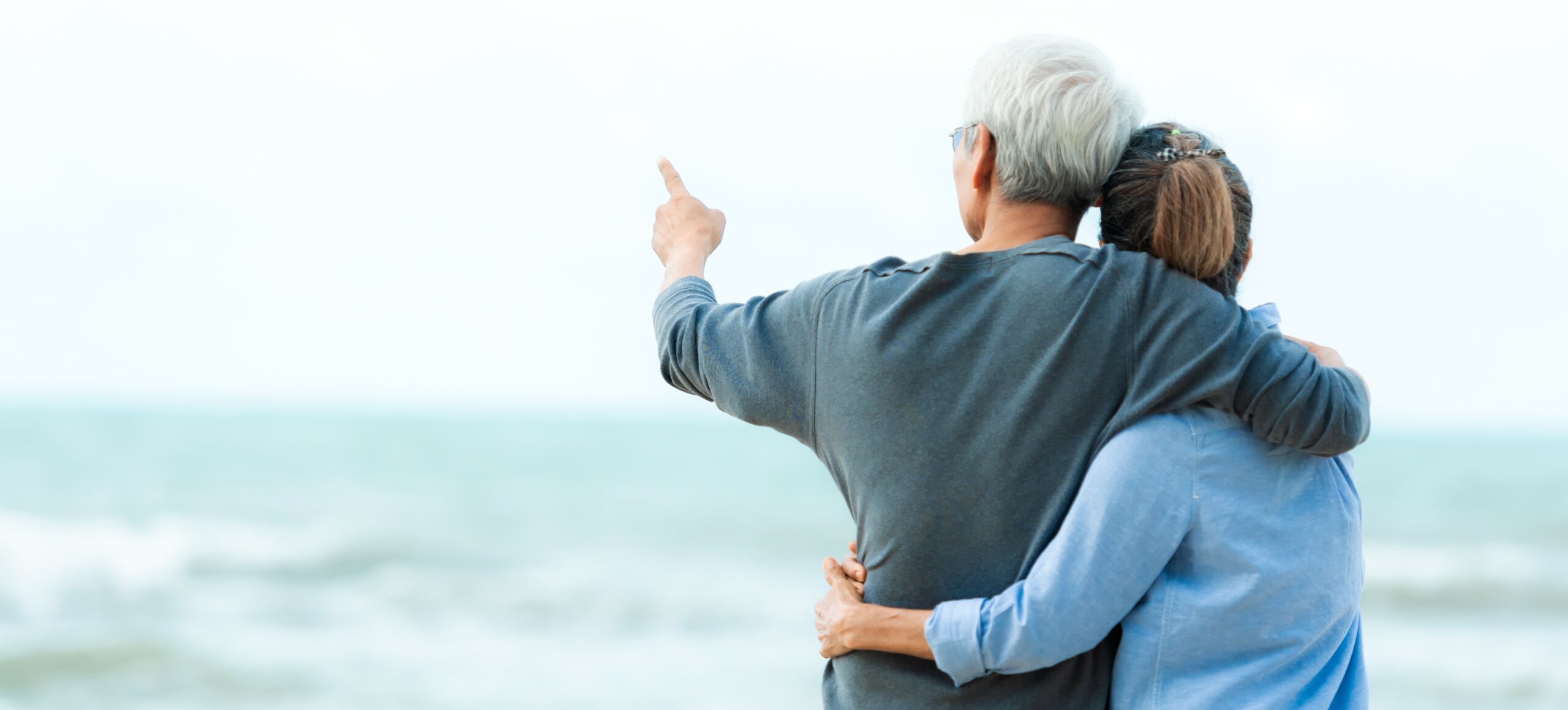 An older couple stands on a beach holding each other and facing away from camera