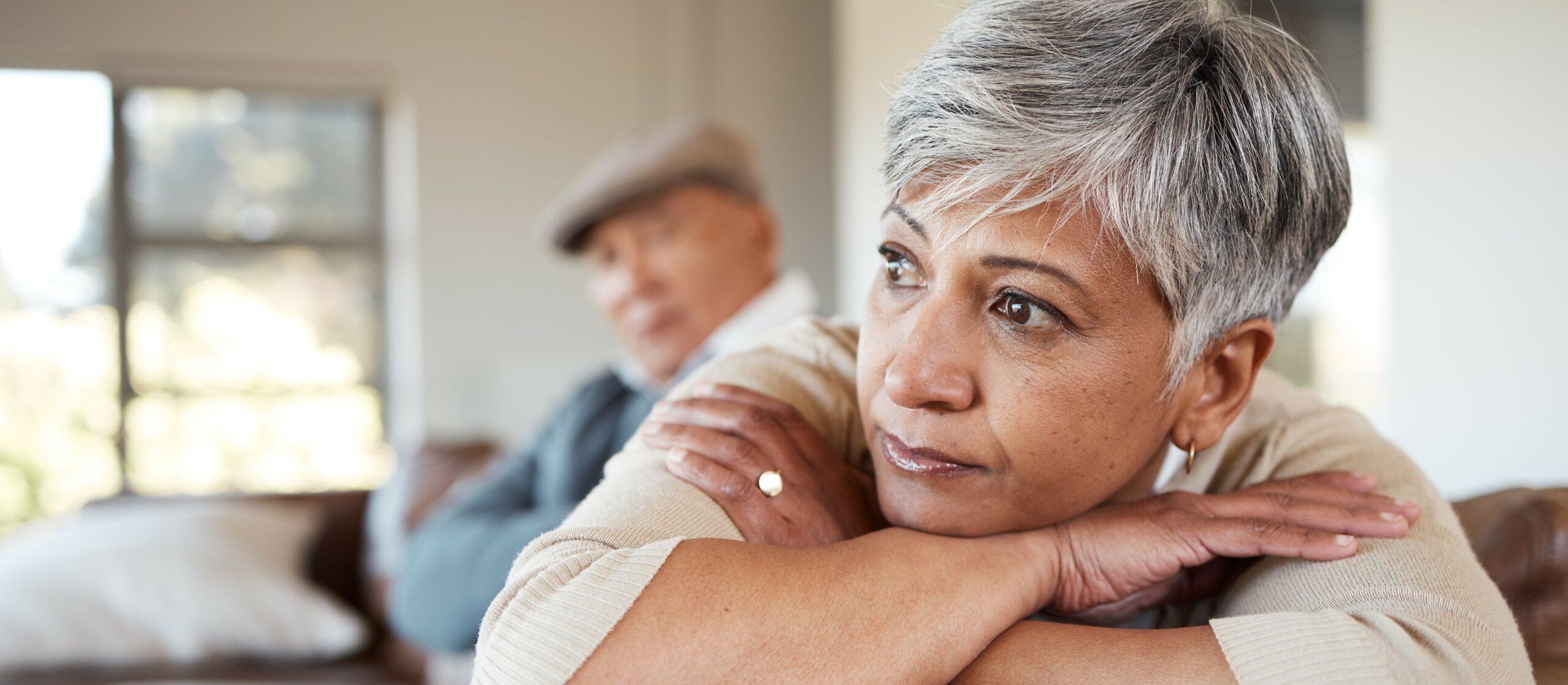 An older woman leans over the arm of a sofa looking sad and thoughtful while her husband sits in the background looking at her with an upset expression