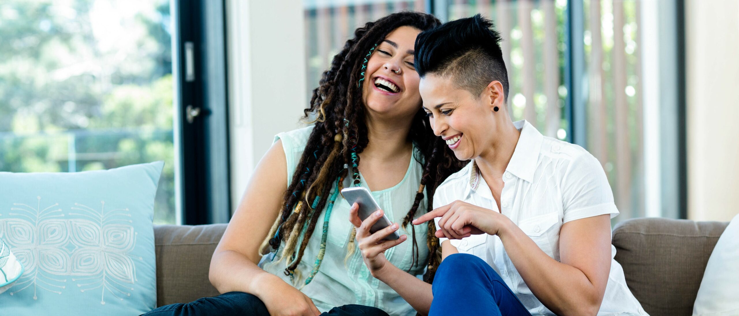 A lesbian couple sits on their couch reading a phone screen, smiling and laughing