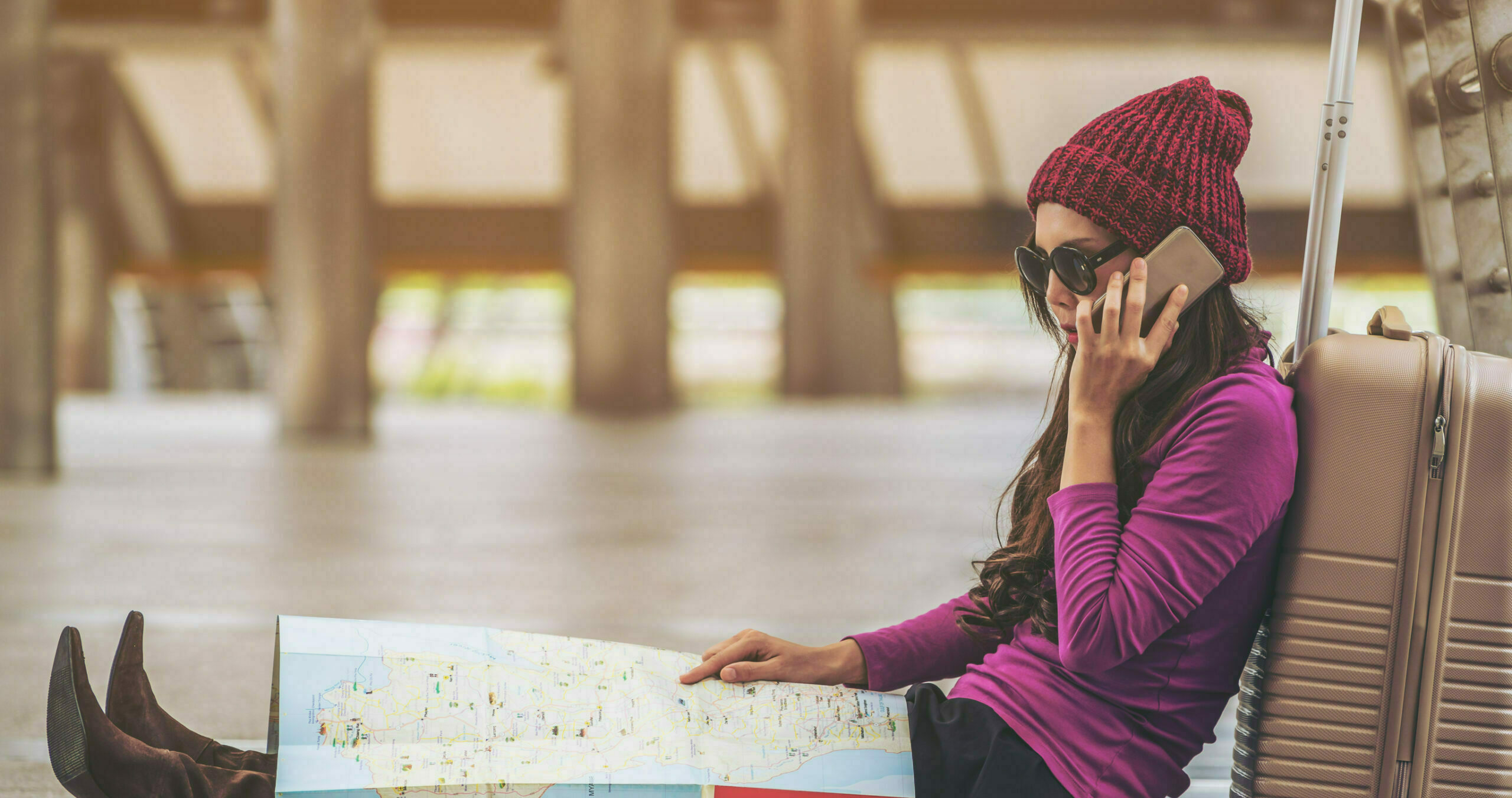 Woman in hat and sunglasses sits on airport floor, leaning against her suitcase and holding a map