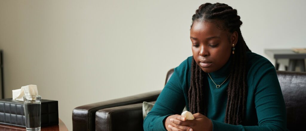 A young Black woman sits on a couch in a therapy office holding a tissue and looking downward while she thinks
